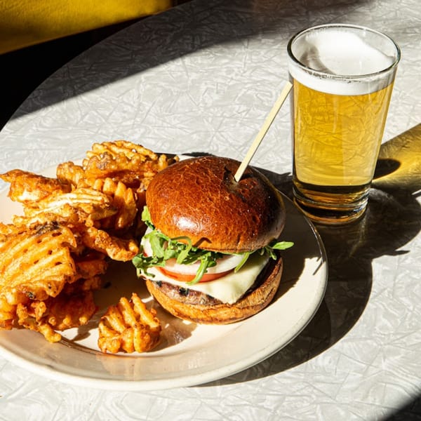Matilda's burger with waffle fries and a glass of beer on a white textured table.