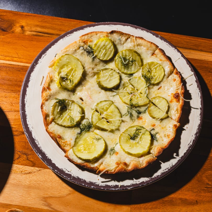 Round flatbread pizza with melted cheese, pickle slices, and fresh dill on a rustic plate.