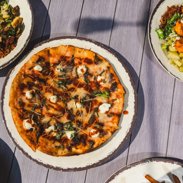 Round pizza with mozzarella, basil, and sauce drizzle on a rustic plate, surrounded by other dishes on a wooden table.