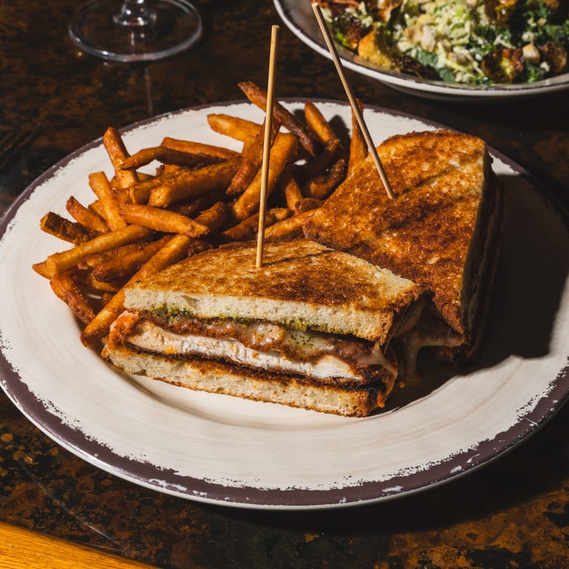 Close-up of a grilled chicken sandwich with melted cheese and pesto, served with french fries.
