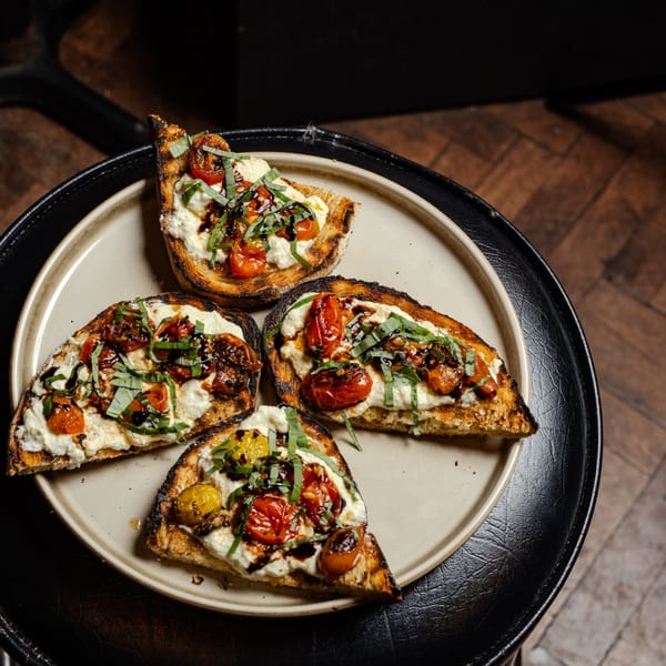 Four grilled toasts with creamy cheese, roasted tomatoes, fresh basil, and balsamic glaze on a plate.