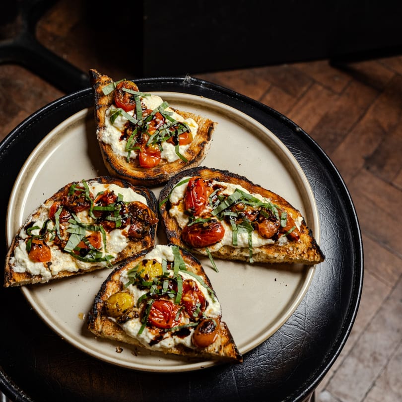 Four grilled toasts with creamy cheese, roasted tomatoes, fresh basil, and balsamic glaze on a plate.