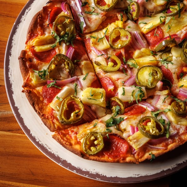 Sliced pizza topped with pepperoni, pineapple, red onion, jalapeños, and fresh herbs.