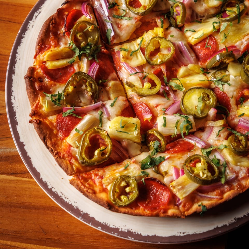 Sliced pizza topped with pepperoni, pineapple, red onion, jalapeños, and fresh herbs.